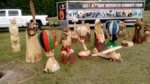 Wooden sculptures of animals and hot air balloons displayed outdoors, with a truck and trailer in the background.