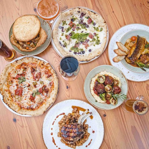 A spread of various dishes including pizzas, meat, and sides on a wooden table, with drinks in glasses.