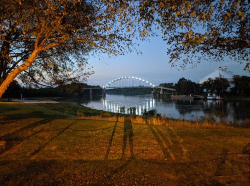 A serene river scene at dusk, featuring a bridge illuminated by lights and shadows cast by nearby trees.