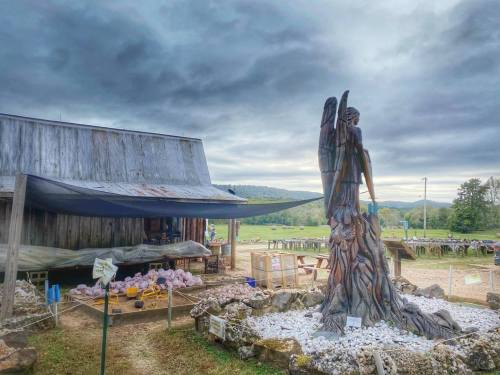 A wooden angel statue stands near a rustic barn, surrounded by rocks and displays of various items under a cloudy sky.