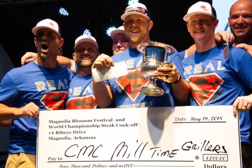 A group of smiling men in blue shirts holding a trophy and a large check at a steak cook-off event.