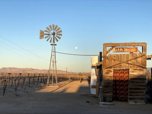 A rustic bathhouse stands beside a windmill under a clear sky with a rising moon in a desert landscape.