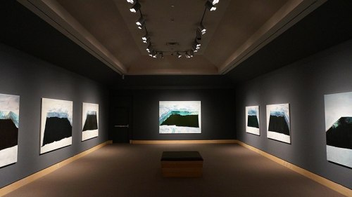 A spacious art gallery featuring large abstract paintings on dark walls, illuminated by overhead lights.