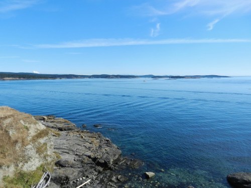 A serene coastal view with calm blue waters, rocky shorelines, and distant islands under a clear blue sky.