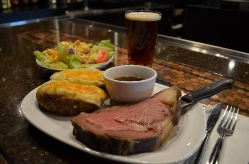 A plate with prime rib, baked potato, gravy, and a side salad, accompanied by a glass of beer on a bar counter.