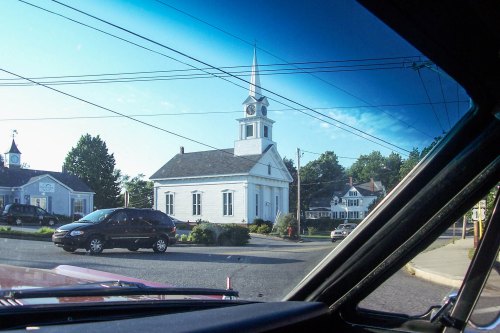View of a white church with a steeple, seen from a car, surrounded by trees and buildings on a sunny day.