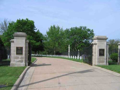 Entrance to a cemetery with stone pillars, green trees, and a pathway lined with white markers.