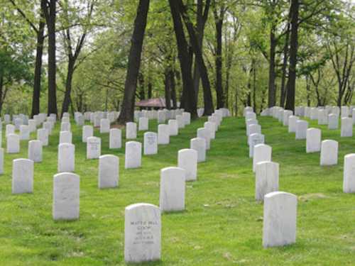 A serene cemetery with rows of white gravestones set in a green, grassy landscape surrounded by trees.