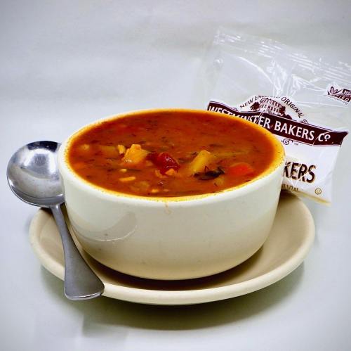 A bowl of hearty soup with vegetables, served with a spoon and a packet of Westminister Bakers crackers.