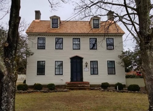 A two-story, white colonial-style house with a sloped roof, surrounded by trees and a grassy lawn.