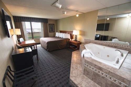 A spacious hotel room featuring a king bed, seating area, desk, and a jacuzzi tub with a large window view.