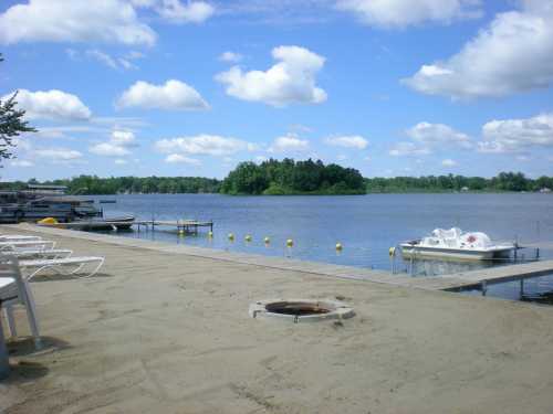 A serene lakeside view with boats, a sandy shore, and fluffy clouds in a blue sky.