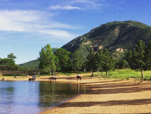 A serene lakeside scene with horses and riders near a sandy shore, surrounded by trees and mountains under a blue sky.