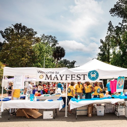 A vibrant outdoor festival scene with tents, people, and a "Welcome to MAYFEST" banner.
