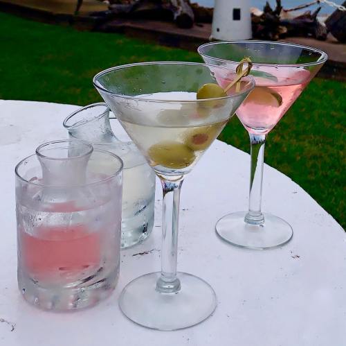 Two martini glasses with olives and a pink drink, alongside a glass of clear liquid, on a table outdoors.