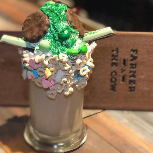 A decadent milkshake topped with green sprinkles, cookies, and cereal, served in a glass with a wooden sign in the background.