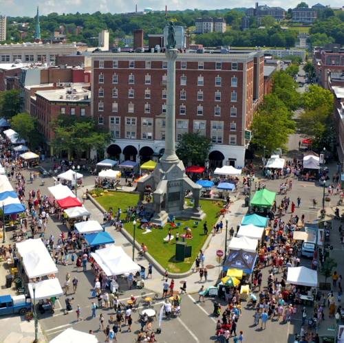 Aerial view of a bustling street fair with tents and crowds around a central monument in a city square.