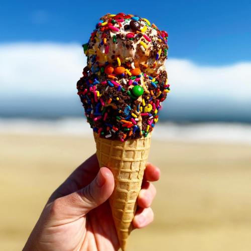 A hand holds a colorful ice cream cone topped with sprinkles and candies against a beach backdrop.