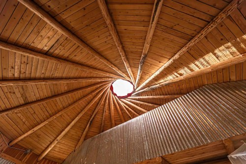 A spiral wooden ceiling with a central circular opening, showcasing beams radiating outward and textured metal walls below.