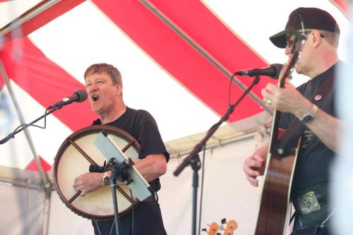 Two musicians perform under a red and white striped tent; one sings passionately while playing a drum, the other strums a guitar.