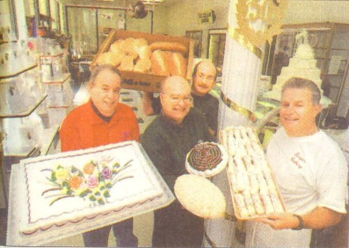 Four men hold various baked goods, including cakes and pastries, in a bakery setting.