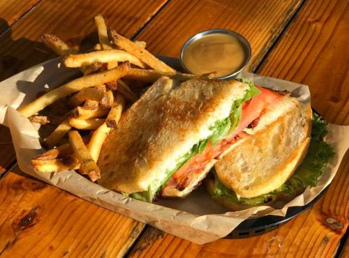A toasted sandwich with lettuce and tomato, served with crispy fries and a small cup of dipping sauce on a wooden table.