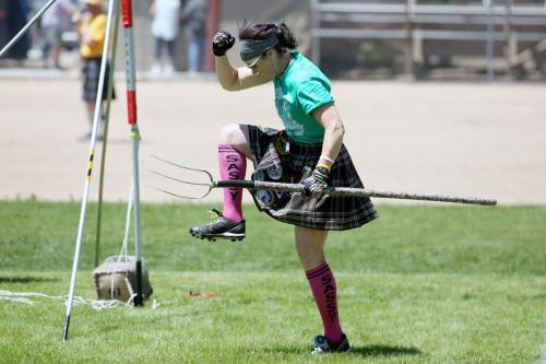 A woman in a kilt and pink socks poses with a pitchfork, raising one leg in a playful stance on a grassy field.