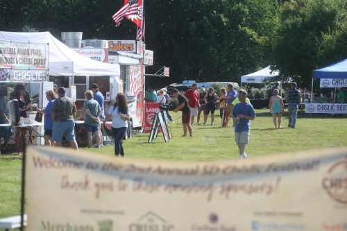 A lively outdoor festival scene with food vendors, people mingling, and a banner welcoming attendees to the Chislic Festival.