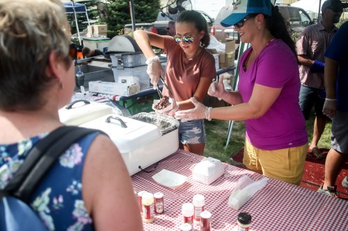 A woman and a girl serve food at a outdoor event, with spices and condiments on a table in front of them.