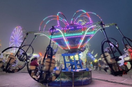 A colorful amusement ride spins in a foggy fairground, surrounded by bright lights and other attractions.