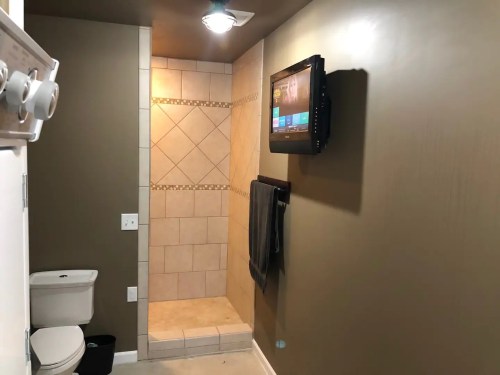 A modern bathroom featuring a shower, toilet, and a wall-mounted TV. Neutral-colored walls and tiled shower area.