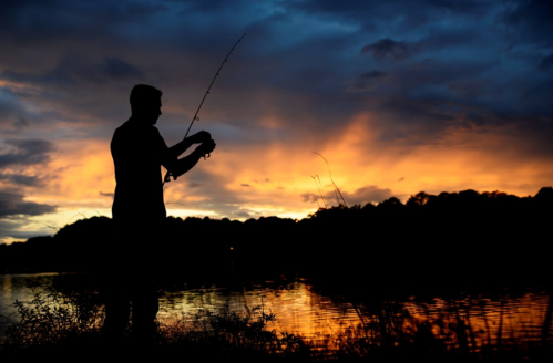 Silhouette of a person fishing at sunset, with vibrant orange and blue skies reflected on the water.