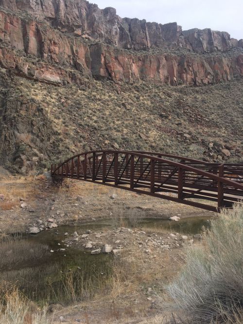 A metal bridge spans a dry riverbed, surrounded by rocky cliffs and sparse vegetation.