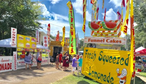 Colorful food stalls at a fair, featuring funnel cakes, lemonade, and BBQ, with people enjoying the festive atmosphere.