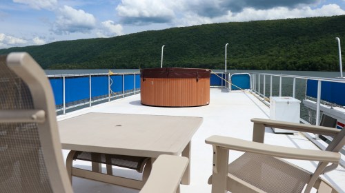 A deck with a hot tub and seating, overlooking a calm river and green hills under a partly cloudy sky.