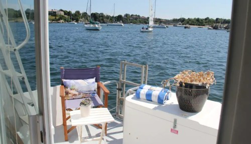 A cozy balcony overlooking a calm waterway with boats, featuring a chair, small table, and potted plant.