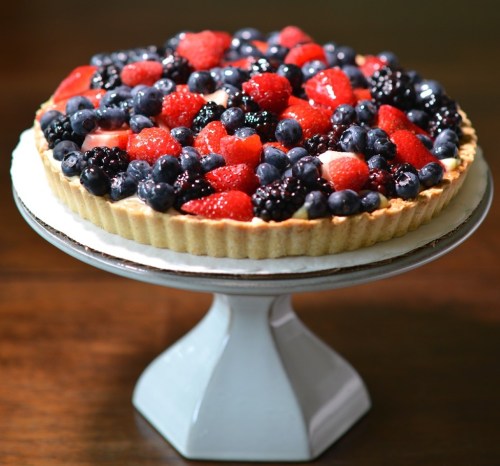 A fruit tart topped with an assortment of fresh berries on a white pedestal stand.