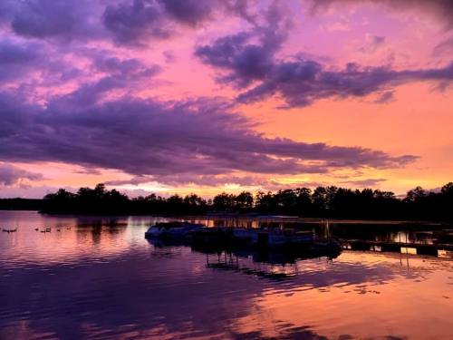 A serene lake at sunset, with vibrant purple and orange skies reflecting on the water and silhouettes of boats.