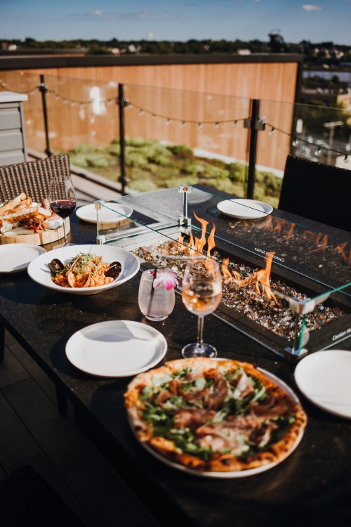 A table set with gourmet dishes, including pizza and pasta, beside a glass fire feature on a sunny outdoor patio.