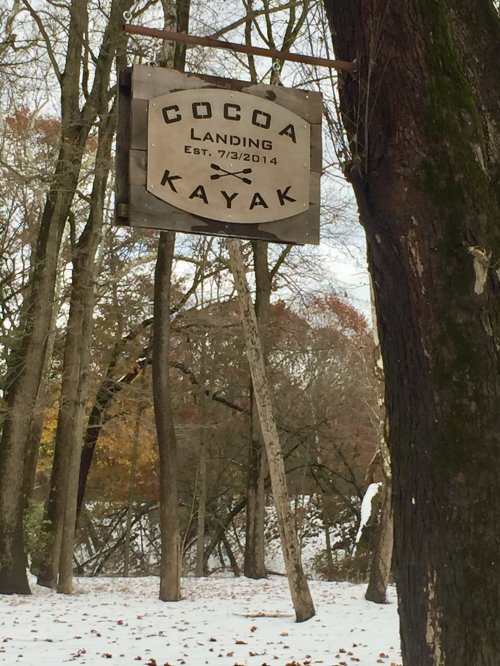 Sign for Cocoa Landing Kayak, established 7/3/2014, surrounded by trees in a snowy landscape.