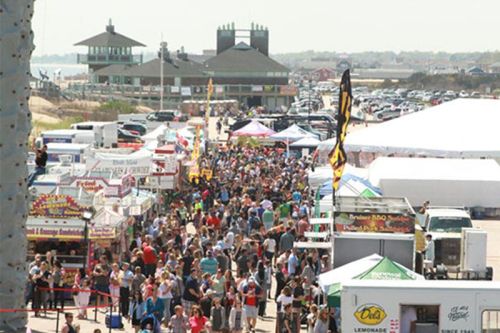 A bustling outdoor festival with crowds, food stalls, and a beachside view in the background.