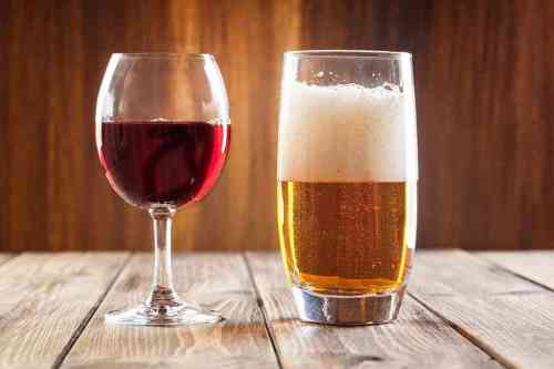 A glass of red wine next to a tall glass of beer, both on a wooden table with a warm background.