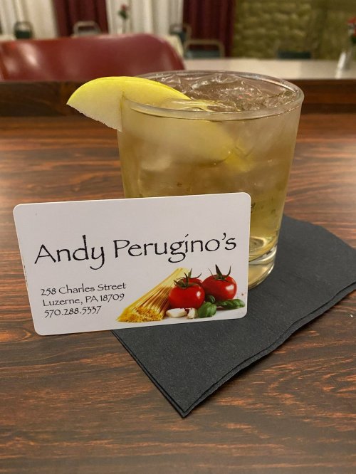 A glass of drink with a lemon slice on a napkin, next to a business card for Andy Perugino's restaurant.