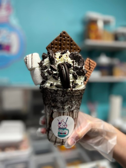 A hand holds a decadent ice cream shake topped with cookies, marshmallows, and chocolate wafers against a colorful background.