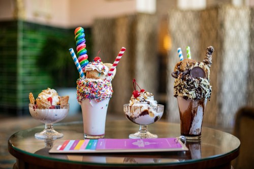 Four colorful milkshakes topped with whipped cream, sprinkles, cherries, and candy, displayed on a table.