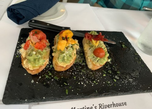 Three colorful crostinis topped with avocado, edible flowers, and herbs on a black slate plate.