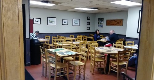 A nearly empty restaurant with wooden tables and chairs, featuring framed artwork on the walls. Two patrons are seated.