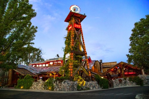 A rustic restaurant with a tall wooden tower, illuminated signs, and greenery, set against a blue evening sky.