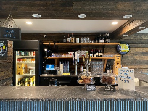 A rustic bar with a wooden shelf displaying drinks, pastries, and a sign inviting customers to place their orders.