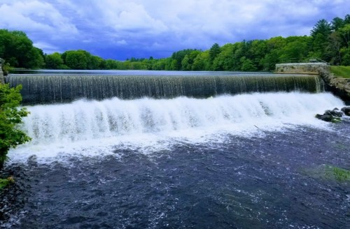 A serene waterfall cascades over a dam, surrounded by lush green trees under a cloudy sky.